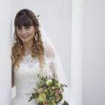 Braut lehnt an weißer Wand mit Brautstrauß